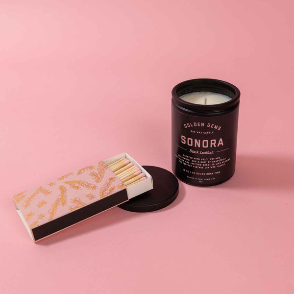 A pink matchbox with a metallic gold design and pink match heads is next to a lit 12 ounce candle in a black jar by Golden Gems