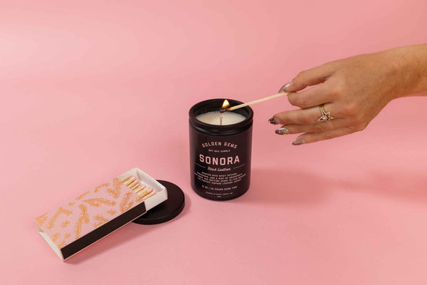 A pink matchbox with a metallic gold design and pink match heads is next to a lit 12 ounce candle in a black jar by Golden Gems