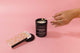 A hand lights a 12 ounce candle in a black jar by Golden Gems and a pink matchbox with a metallic gold design sits next to the candle