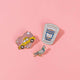 Set of three glittery enamel pins, a pink heart with a yellow taxi, a small pigeon, and a coffee cup that reads Caffeine Addict