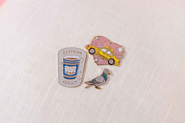 Set of three glittery enamel pins, a pink heart with a yellow taxi, a small pigeon, and a coffee cup that reads Caffeine Addict