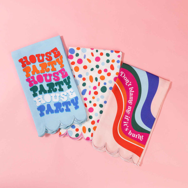 Set of three brightly colored tea towels with scalloped edges each with a different design, a light blue towel with a groovy font repeatedly reads house party, a white towel with multi-colored abstract dots, and a blush pink towel with a multi-colored wave design